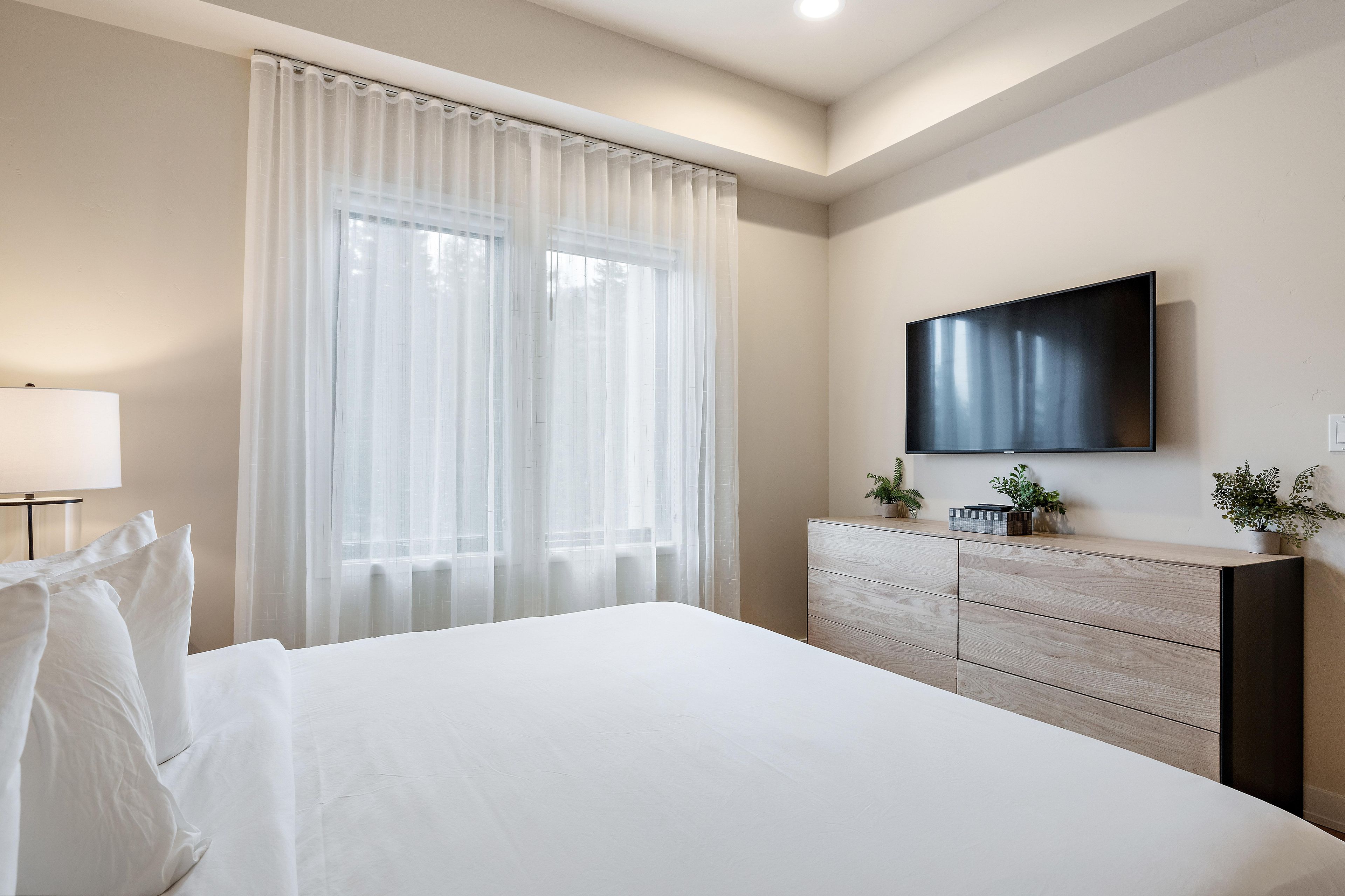 Hotellihuone, jossa on sänky, lipasto, seinälle asennettu televisio ja läpikuultavat verhot ikkunoissa.