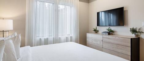 Hotellihuone, jossa on sänky, lipasto, seinälle asennettu televisio ja läpikuultavat verhot ikkunoissa.