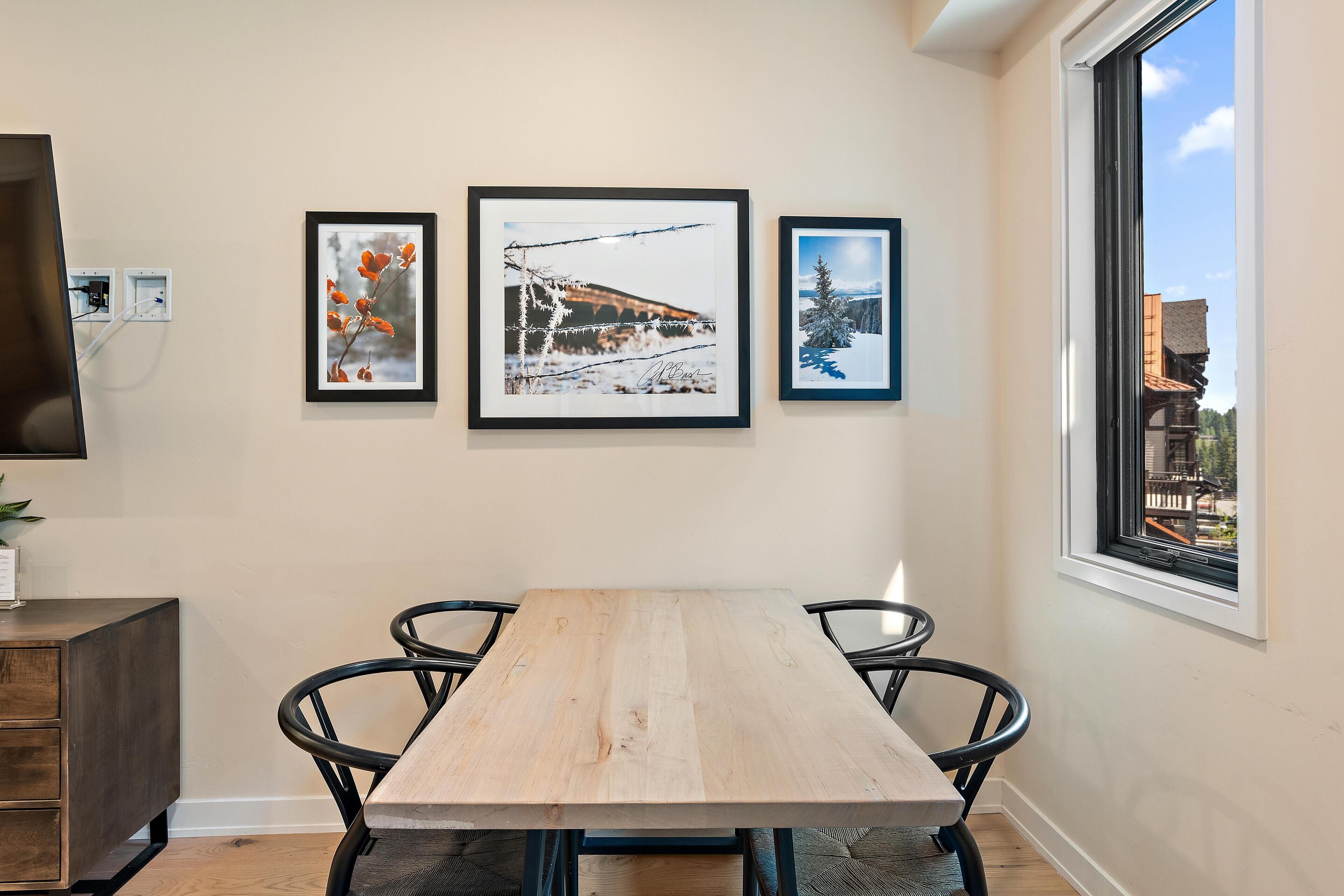 A dining area with a wooden table and black chairs, framed pictures on the wall, and a window with a view of buildings.