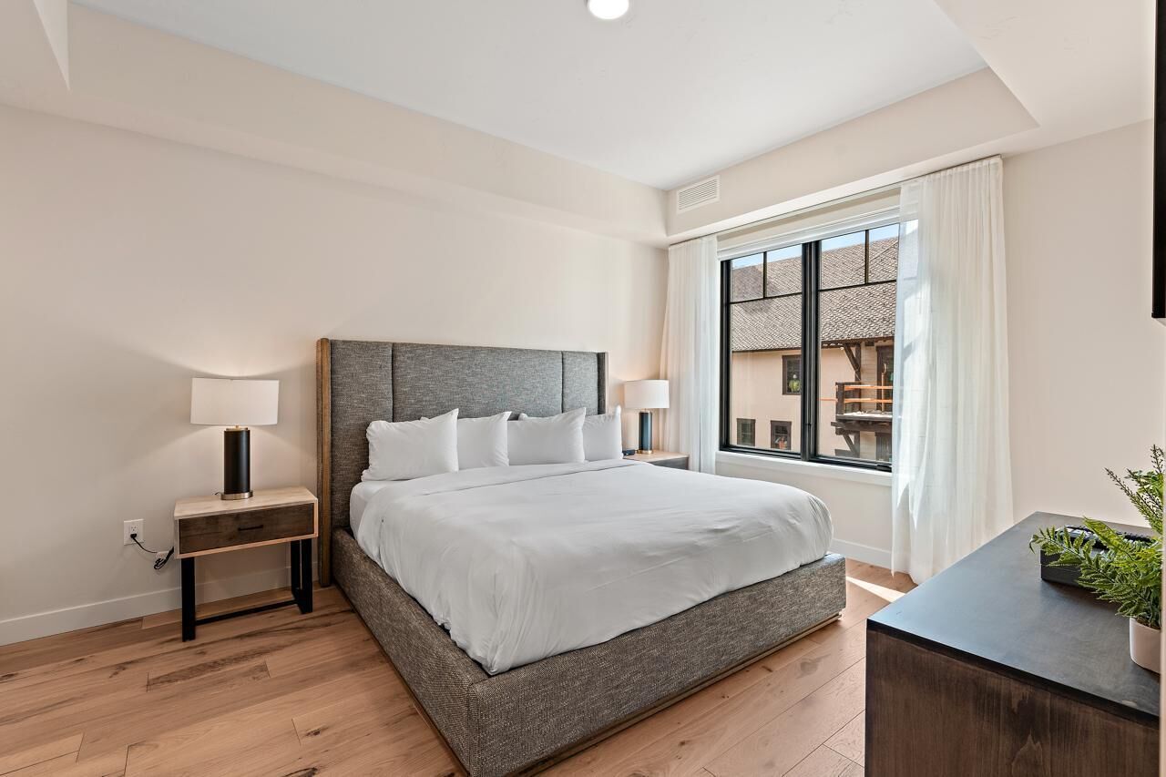 A modern bedroom with a large bed, bedside tables with lamps, a window with curtains, and a view of the outside.