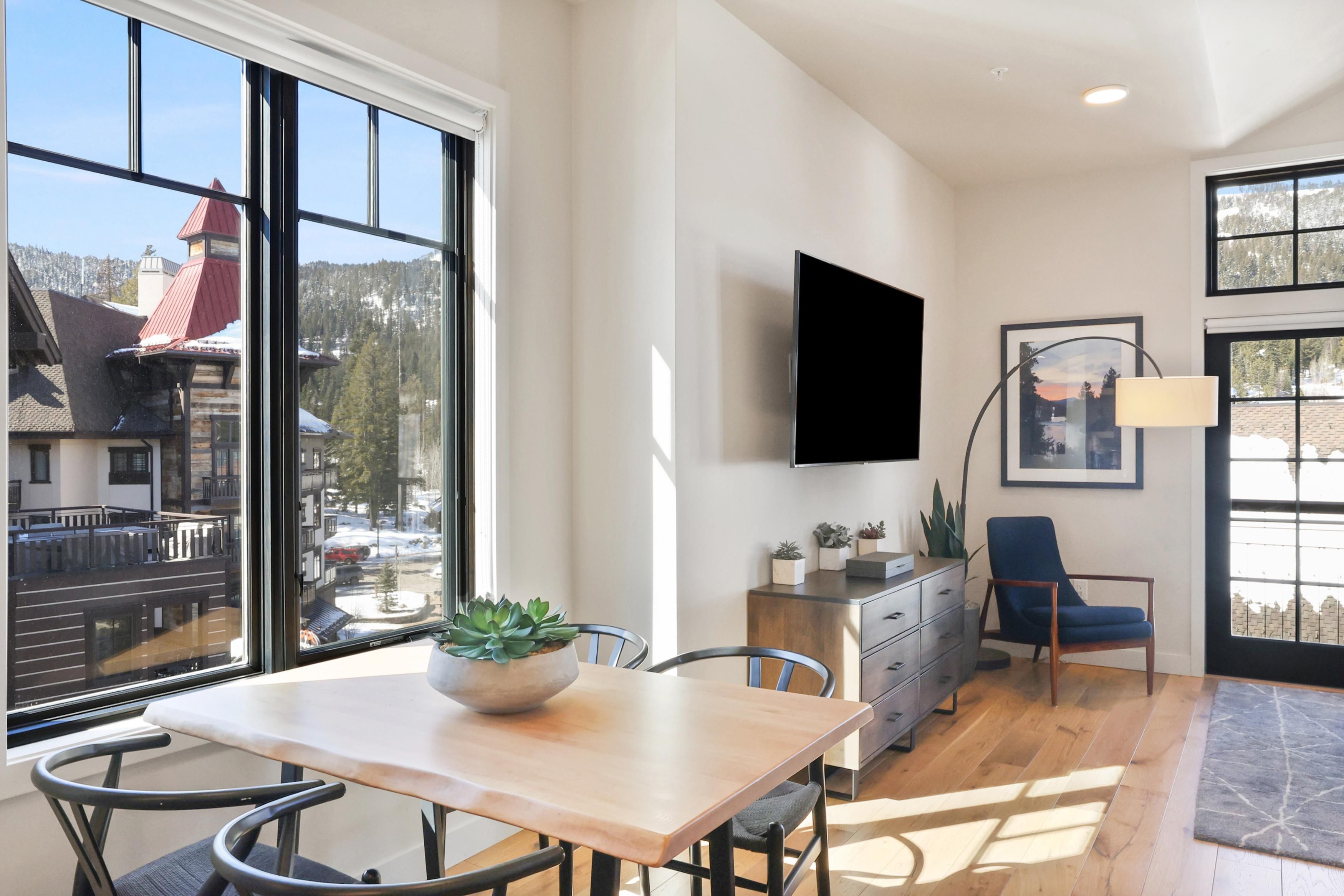 Una sala moderna con un amplio ventanal, un televisor de pantalla plana montado en la pared, una mesa de comedor con sillas y un sillón azul.