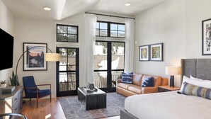Une chambre d’hôtel moderne équipée d’un lit, d’un canapé, d’un fauteuil, d’un bureau et d’une grande fenêtre donnant sur des arbres et des immeubles.