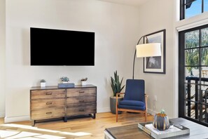 Een moderne woonkamer met een flatscreen televisie aan de muur, een houten kast met lades, een blauwe fauteuil en een vloerlamp.