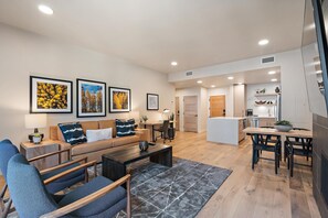 Una sala de estar moderna con sofá, mesa de centro y sillones, un área de cocina con mesa de comedor, y un pasillo que lleva a un dormitorio.