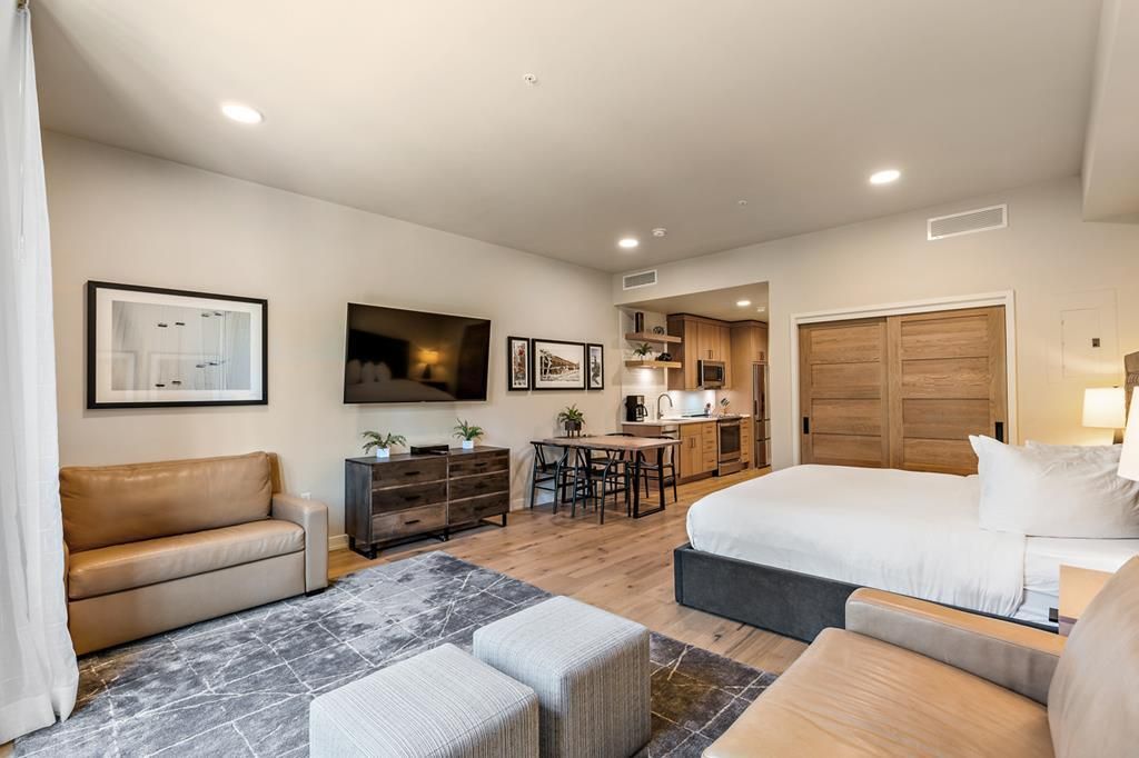 A modern hotel room with a large bed, a sofa, a desk, and a kitchenette.
