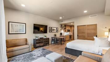 A modern hotel room with a large bed, a sofa, a desk, and a kitchenette.