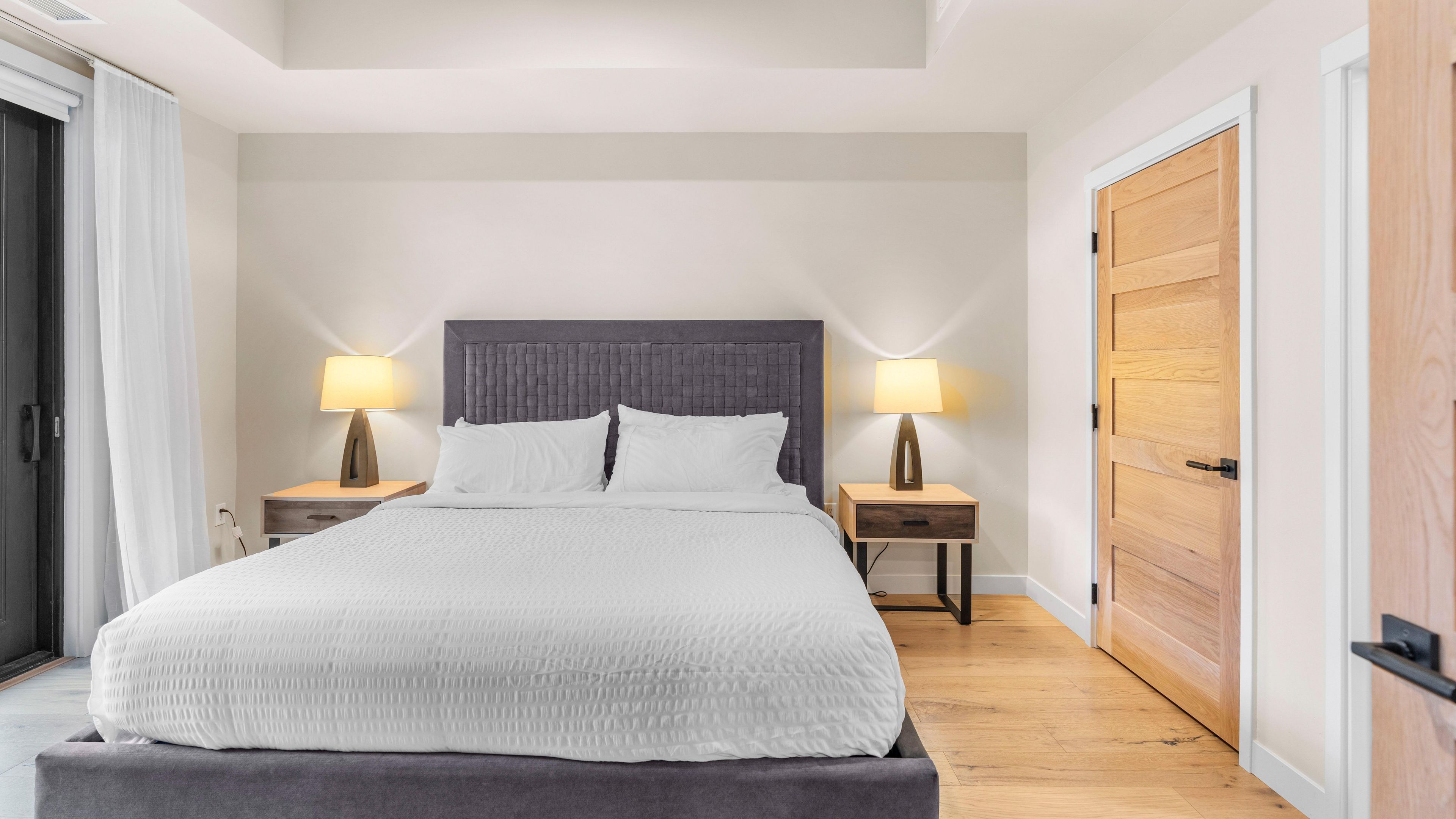 Una cama bien hecha con cabecera gris, dos mesitas de noche con lámparas y una puerta de madera.