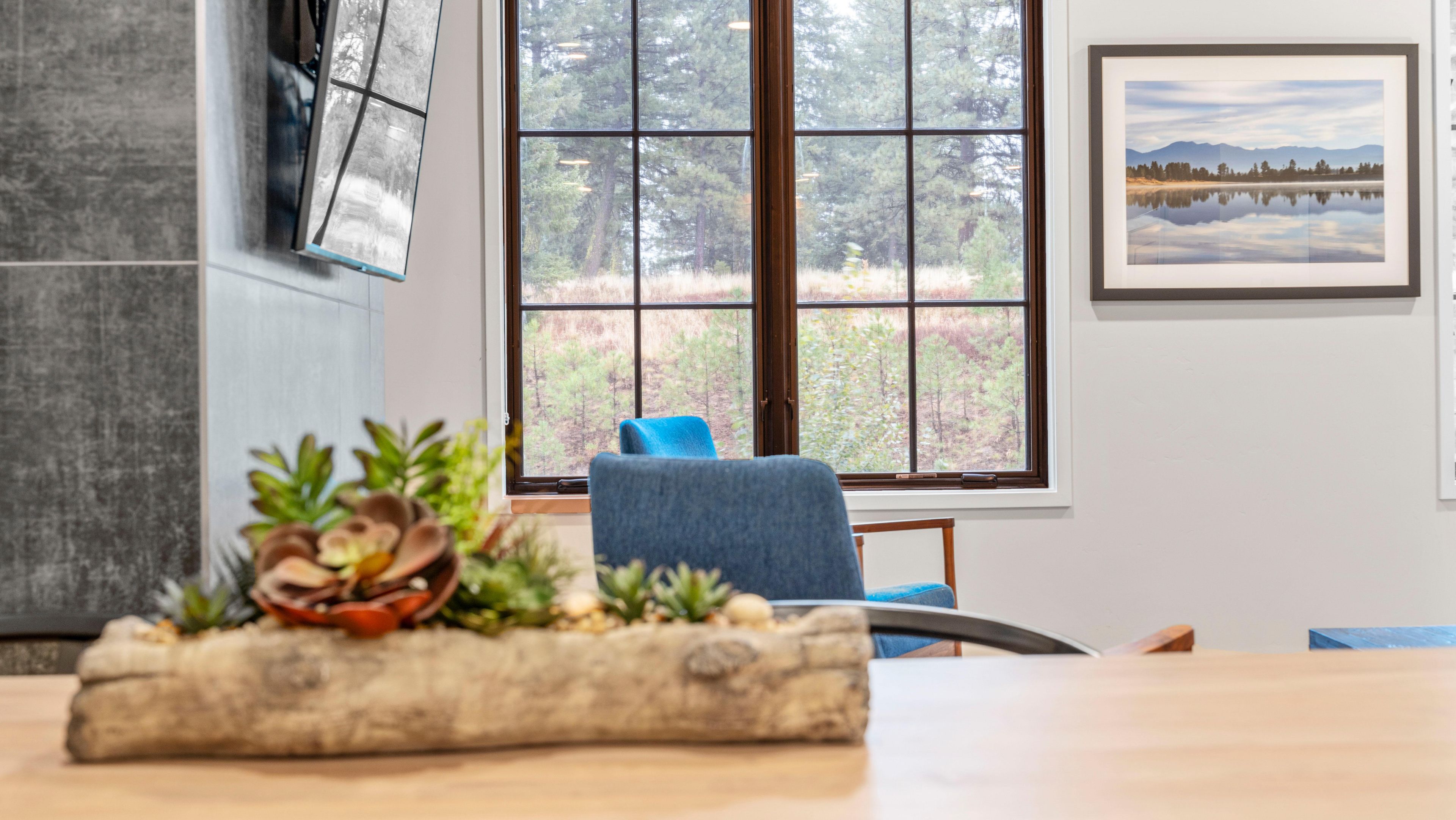 A room with a large window, a blue chair, a table with a decorative plant, and a framed picture on the wall.