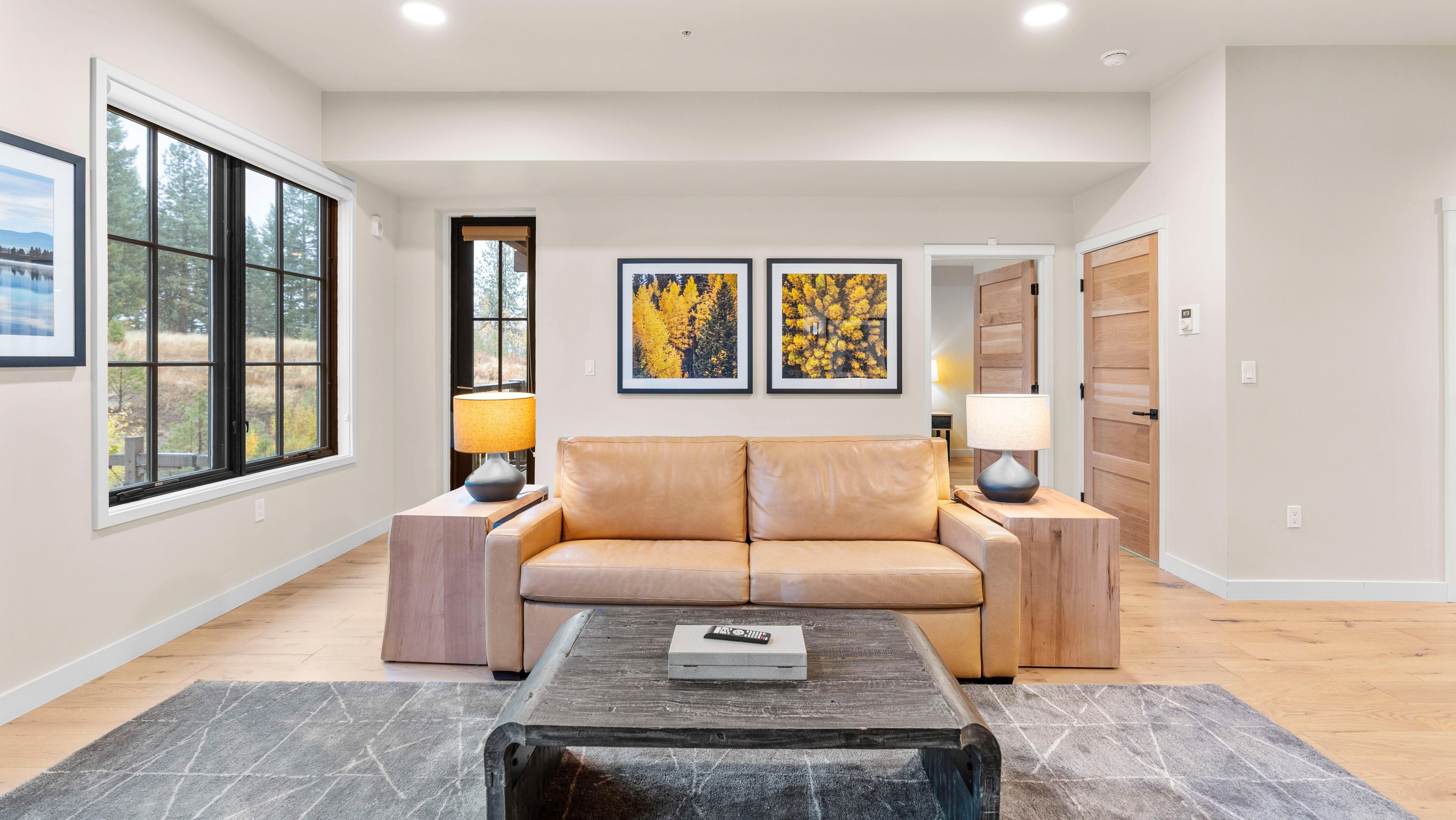 A modern living room with a leather sofa, a coffee table, and two lamps. There are large windows and framed artwork on the walls.