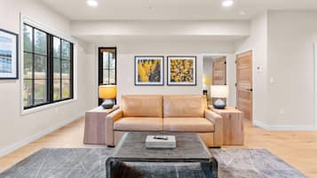 A modern living room with a leather sofa, a coffee table, and two lamps. There are large windows and framed artwork on the walls.
