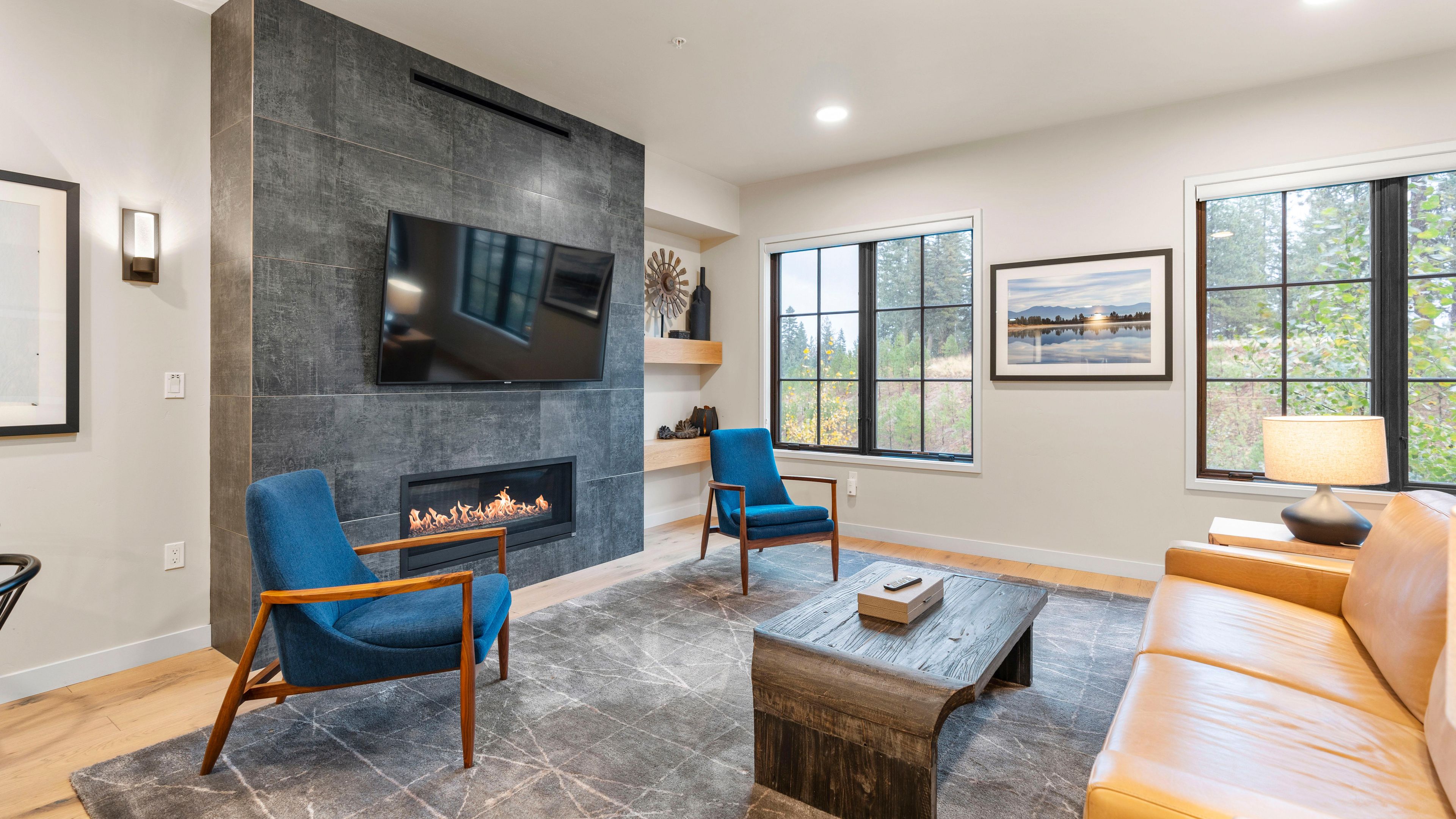 A modern living room with a fireplace, a flat-screen TV, and a large window with a view of trees.
