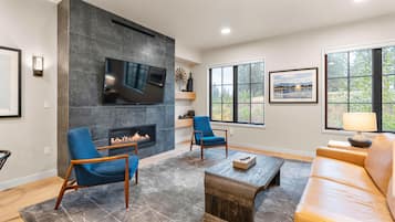 A modern living room with a fireplace, a flat-screen TV, and a large window with a view of trees.