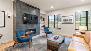 A modern living room with a fireplace, a flat-screen TV, and a large window with a view of trees. - Mountain Condo | Includes Two 1-Day Lift Tickets (Tamarack)
