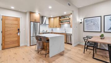 A modern kitchen with wooden cabinets, a stainless steel refrigerator, a white marble island, and a dining area with a table and chairs.