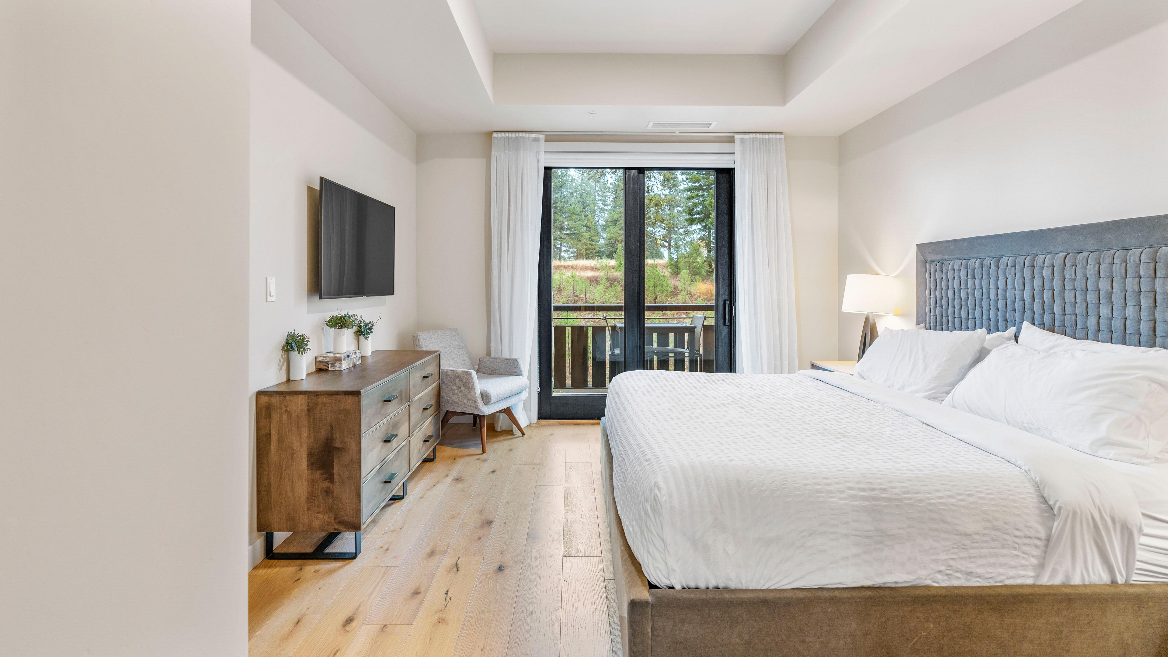 A hotel room with a bed, a TV mounted on the wall, a desk with a chair, and a balcony with a view of trees.