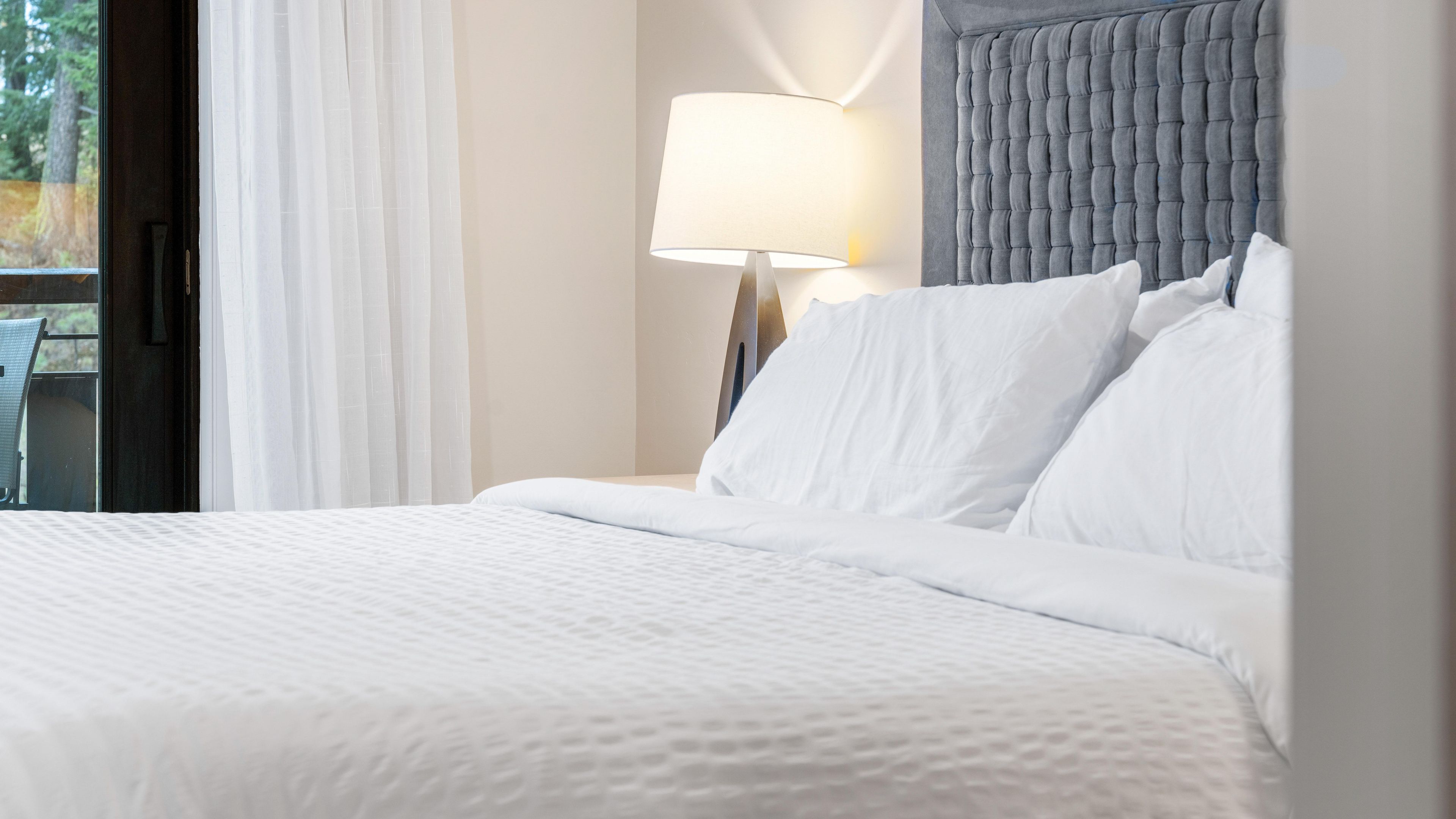 Una cama bien hecha con ropa blanca, cabecera tapizada de gris y una lámpara de noche.