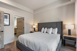 A modern hotel room with a large bed, two bedside tables with lamps, a wooden door, and a framed picture on the wall.