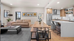 Una sala de estar moderna con una mesa de comedor de madera, un sofá y una zona de cocina con una isla grande y taburetes de bar.