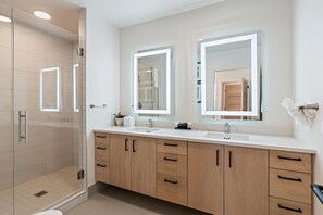 A modern bathroom with a glass shower enclosure, a double sink vanity with wooden cabinets, and two large mirrors with integrated lighting.