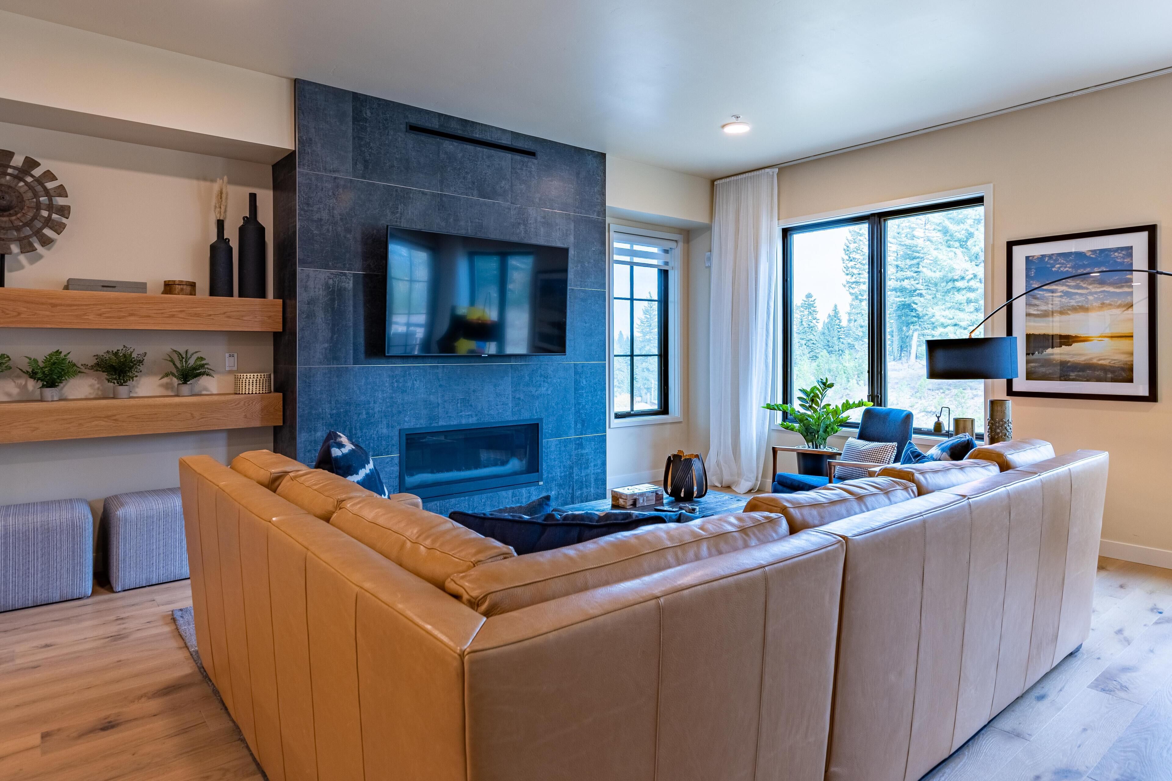 A modern living room with a sectional sofa, a fireplace, and a large window providing a view of trees.