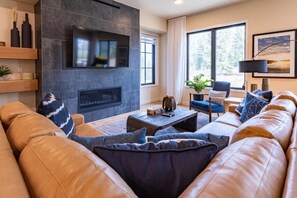 A modern living room with a leather sofa, a fireplace, a television, and a large window with a view of trees.
