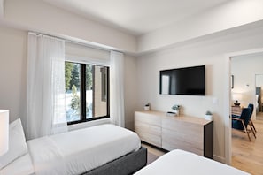 A modern hotel room with a large bed, a desk with a flat-screen TV, a dresser, and a view of the outdoors.