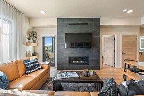 A modern living room with a fireplace, a sofa, a coffee table, and a dining area. - Scarlet Tanager | Includes Two 1-Day Lift Tickets (Tamarack)
