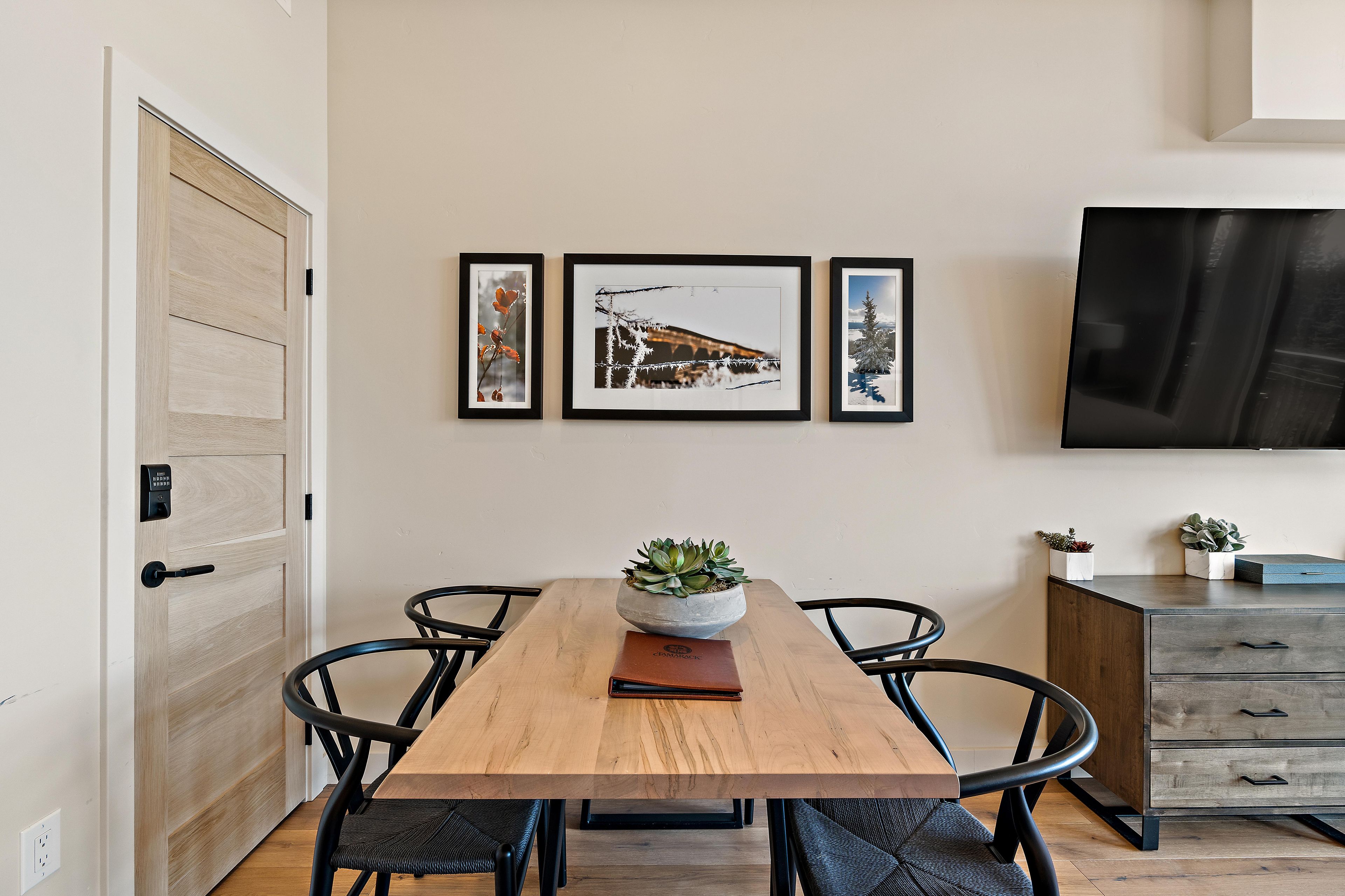 A dining area with a wooden table, black chairs, a potted plant, and a TV mounted on the wall.