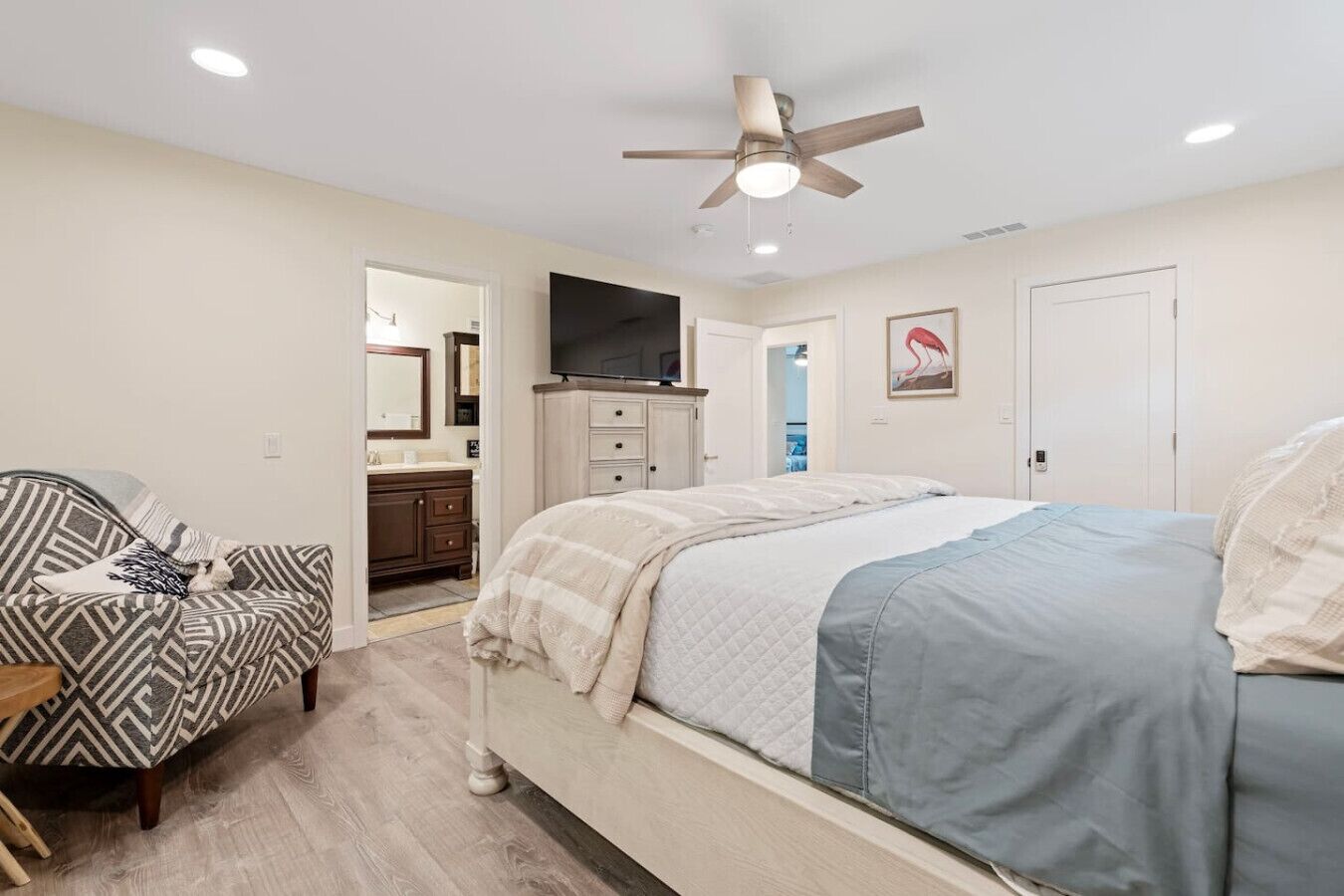 A bedroom with a bed, a dresser, a vanity, a ceiling fan, and a door leading to another room.