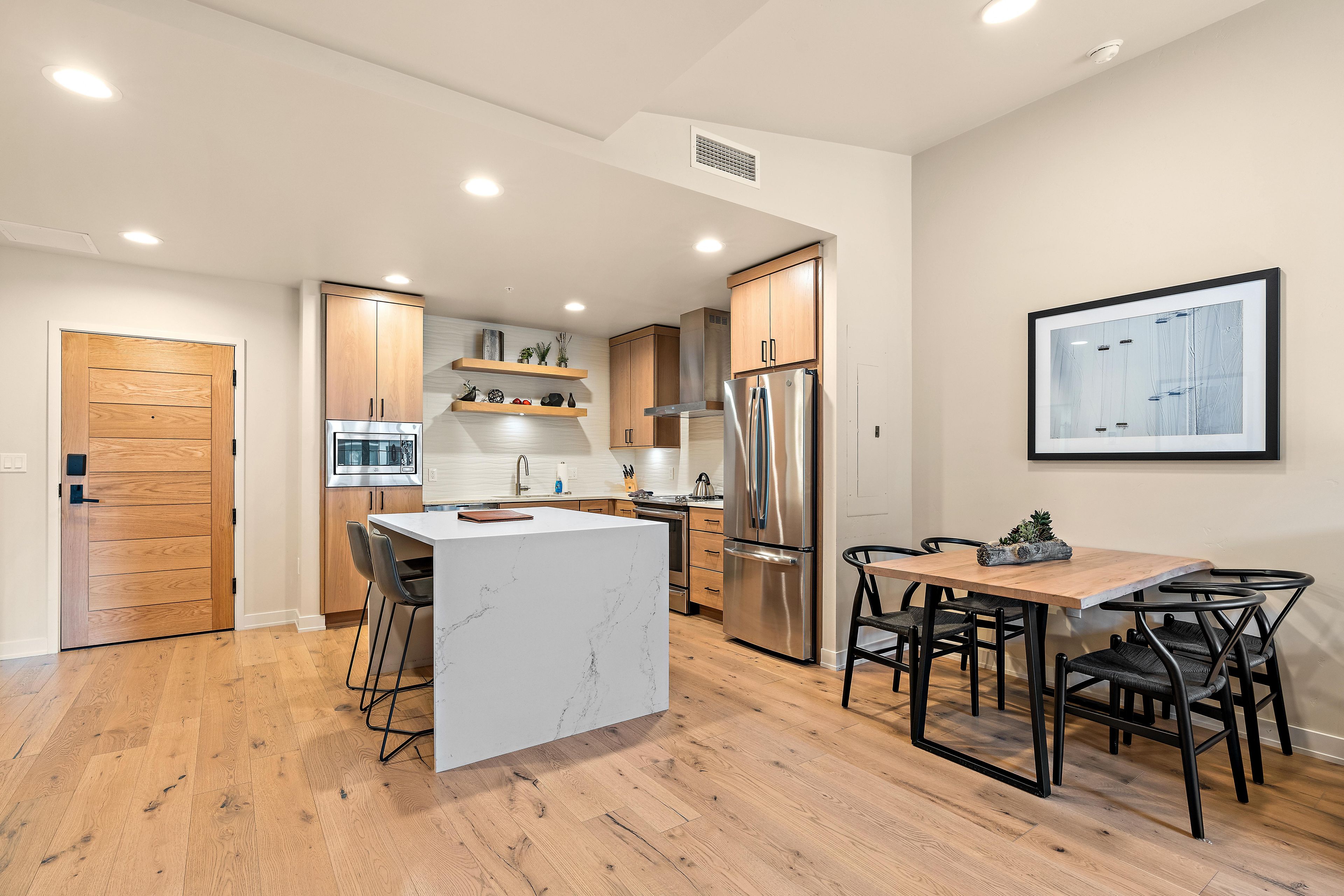 A modern kitchen with a central island, wooden cabinets, stainless steel appliances, and a dining area with black chairs.