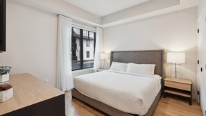 Habitación de hotel con cama, lámparas de noche, mesita de noche, ventana con cortinas y un escritorio de madera con una planta.