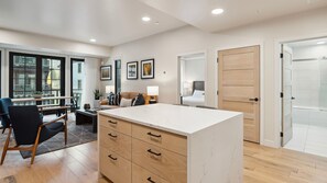 Une chambre d’hôtel moderne dotée d’un grand comptoir central, d’un coin salon avec un canapé et des fauteuils, d’une table à manger et offrant une vue sur la ville grâce à de grandes fenêtres.