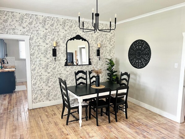 A dining area with a black table, four chairs, a mirror, a chandelier, and a wall with floral wallpaper.