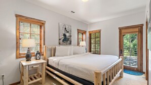 A bedroom with a wooden bed, two bedside tables with lamps, a large window with blinds, and a door leading to an outdoor area. - Lone Peak | Includes Two 1-Day Lift Tickets (Tamarack)