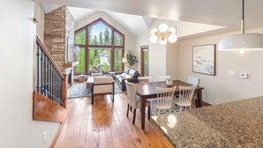 A spacious living area with a high ceiling, wooden flooring, a stone accent wall, a dining table with chairs, and a large window offering a view of greenery. - Aspen Grove | Includes Two 1-Day Lift Tickets (Tamarack)