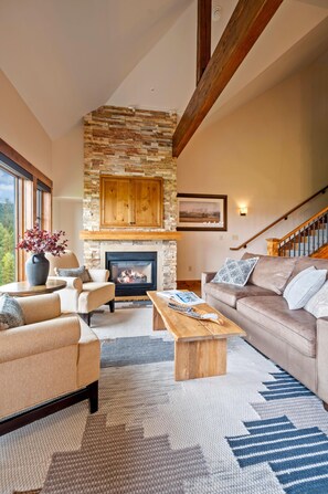 A living room with a stone fireplace, wooden beams, a sofa, a coffee table, and a view of the outdoors. - Lake View Retreat|Exclusive Tamarack Perks (Tamarack)
