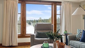 Ein Zimmer mit großem Fenster, einem grauen Sessel, einem Holztisch und einer Couch mit gemusterten Kissen.