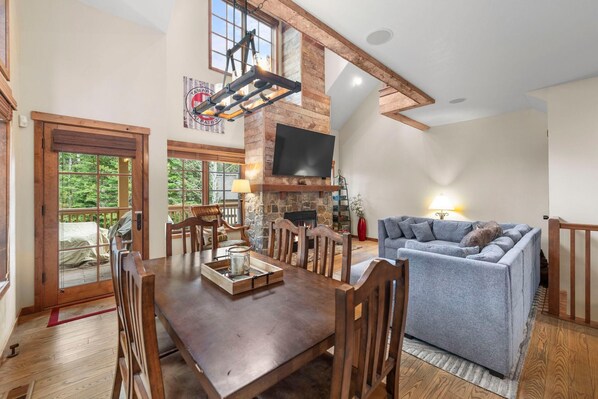 A spacious living room with a wooden dining table, a fireplace, and a television mounted on the wall. - Base Camp | Includes Two 1-Day Lift Tickets (Tamarack)
