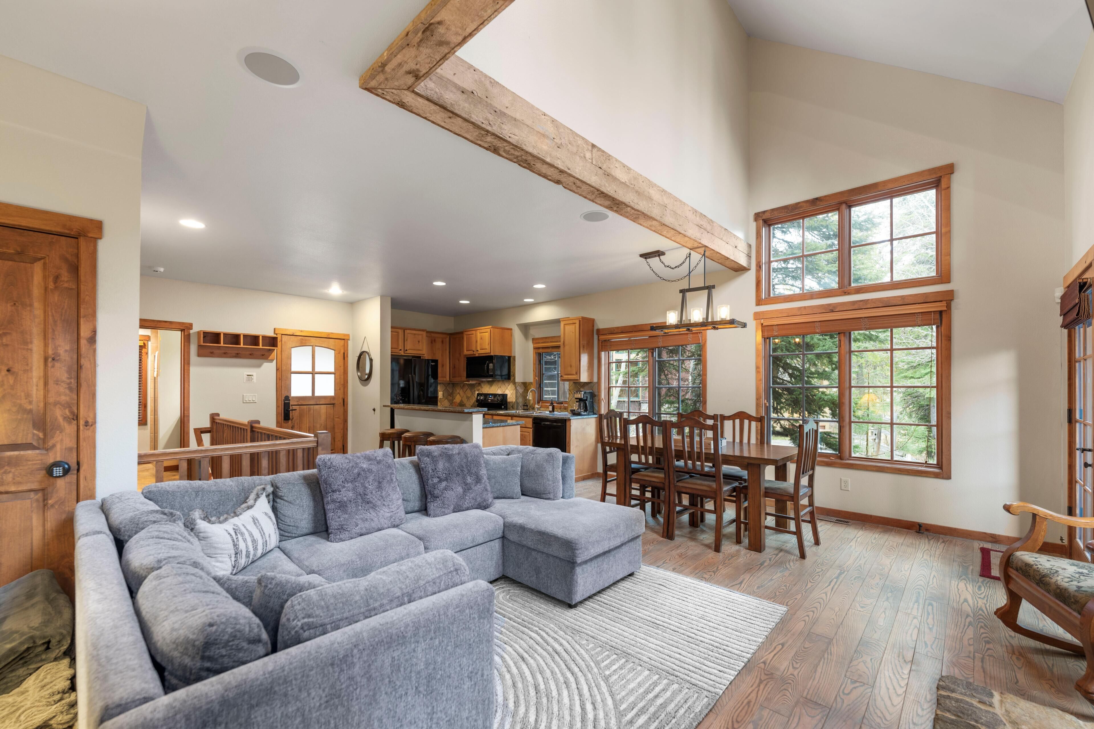Amplia sala de estar con sofá seccional gris, mesa de comedor de madera y sillas, cocina con armarios de madera, y ventanas grandes que ofrecen vistas al exterior.