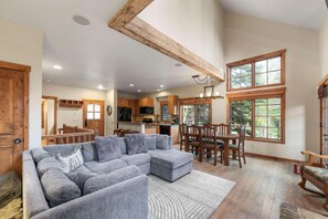 A spacious living area with a gray sectional sofa, wooden dining table, and chairs, a kitchen with wooden cabinets, and large windows offering views of the outdoors. - Base Camp | Exclusive Tamarack Perks (Tamarack)