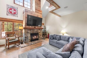 A living room with a stone fireplace, a flat-screen TV, a grey sofa, and a wooden rocking chair. - Base Camp | Exclusive Tamarack Perks (Tamarack)