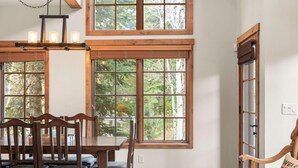 A dining area with a wooden table and chairs, large windows with a view of trees, and a hanging light fixture. - Base Camp | Includes Two 1-Day Lift Tickets (Tamarack)