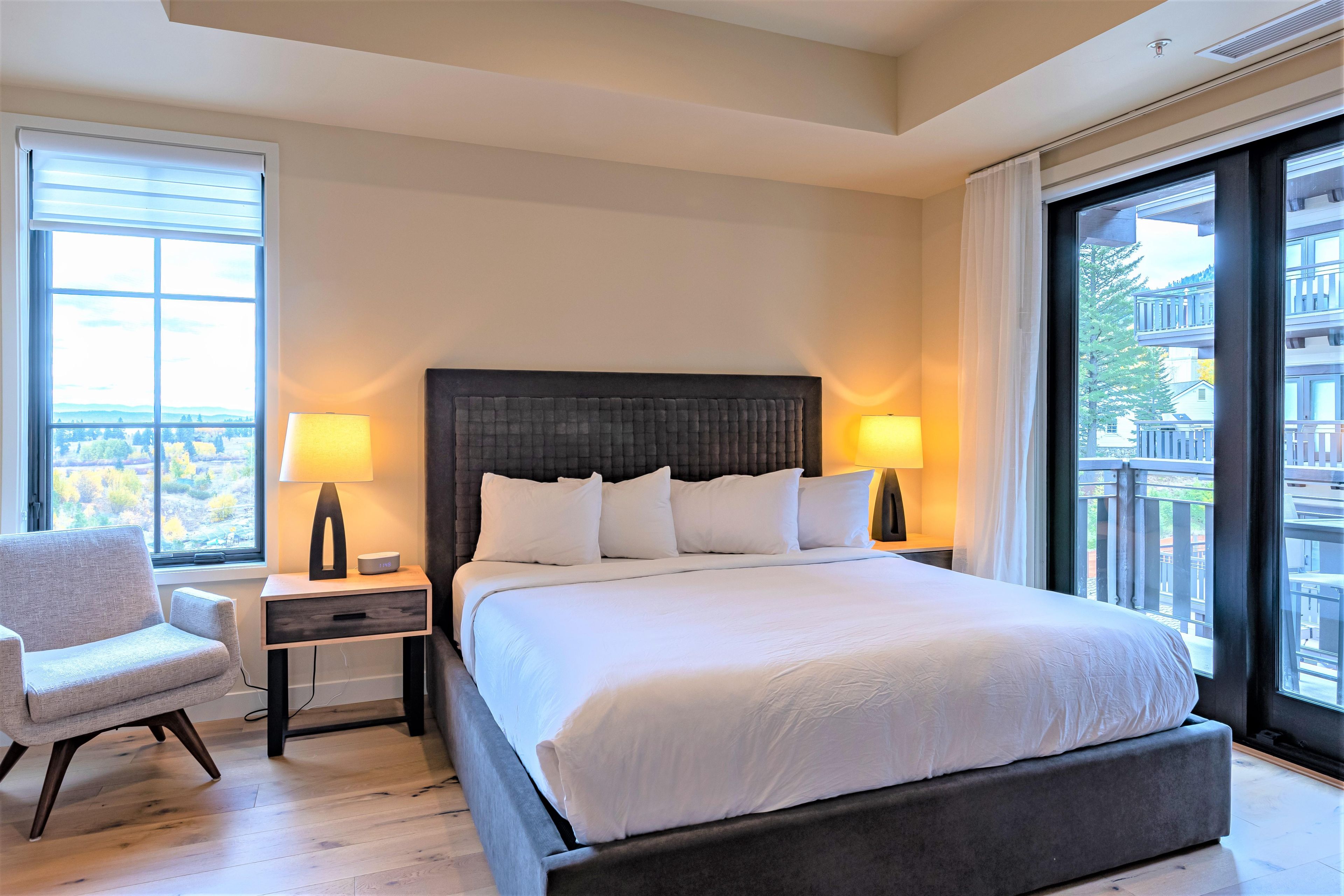 A modern hotel room with a large bed, two bedside tables with lamps, a chair, and a view of a cityscape through large windows.