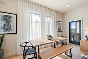 Un comedor con mesa de madera, sillas, un banco, una planta en maceta y vistas a través de un ventanal.