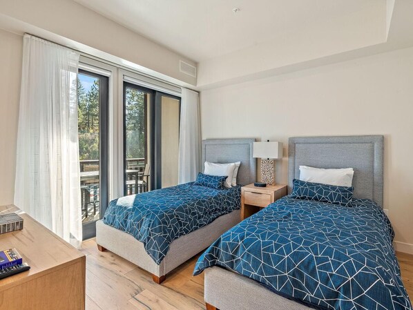 A hotel room with two beds, a wooden desk, a nightstand, and a sliding glass door leading to an outdoor area.