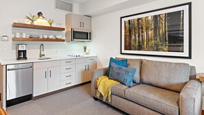 Cocina moderna con armarios blancos, microondas y cafetera. Hay un sofá gris con una manta amarilla y almohadas azules. Un cuadro de un bosque enmarcado está en la pared.
