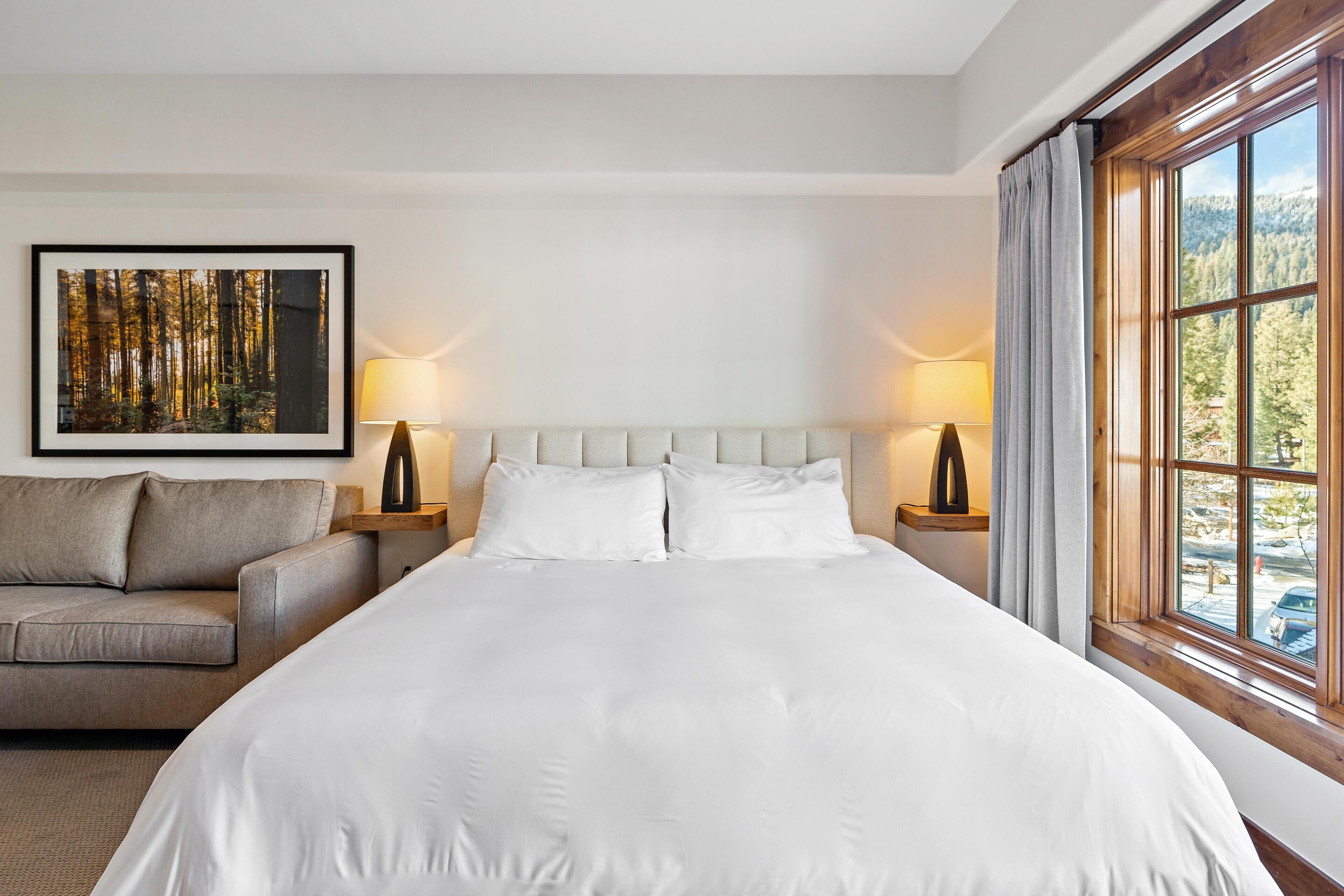 A hotel room with a large bed, two bedside lamps, a sofa, a framed picture on the wall, and a window with a view of trees.