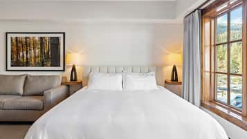 A hotel room with a large bed, two bedside lamps, a sofa, a framed picture on the wall, and a window with a view of trees.