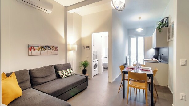 A modern living room with a grey sofa, a dining table with chairs, and a kitchen area in the background. - Luci A Lago (Como)