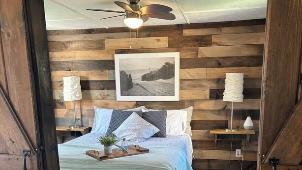 A bedroom with a wooden wall, a bed with a striped bedspread, two bedside lamps, a ceiling fan, and a framed picture on the wall.